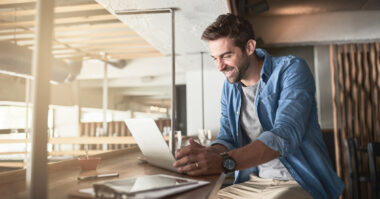 A man sits at a wooden counter in a bright, modern space, using a laptop and smiling—perfect for entrepreneurs. Papers, a pen, and a coffee cup are scattered on the counter beside him.