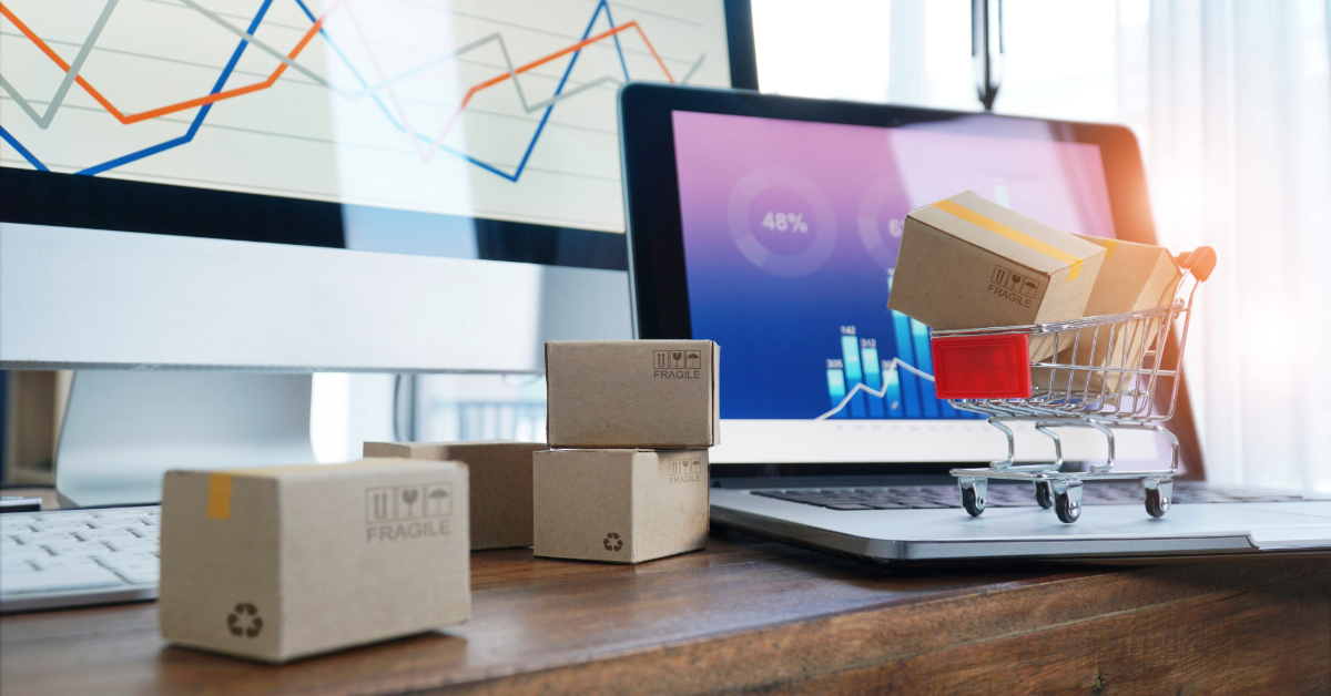 Small cardboard boxes and a miniature shopping cart sit on a desk with a laptop and monitor displaying charts, illustrating strategies for ecommerce success in online shopping and digital business.