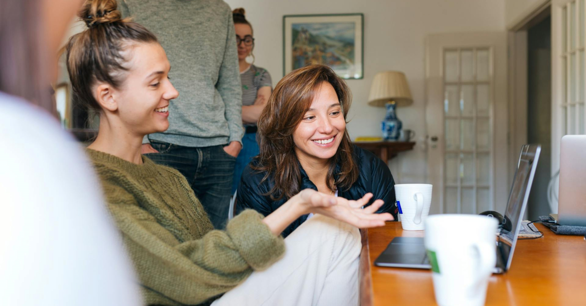 Two women sit at a table, smiling and talking while looking at a laptop, sharing proven tips. Three other people stand in the background in a bright, casual room.