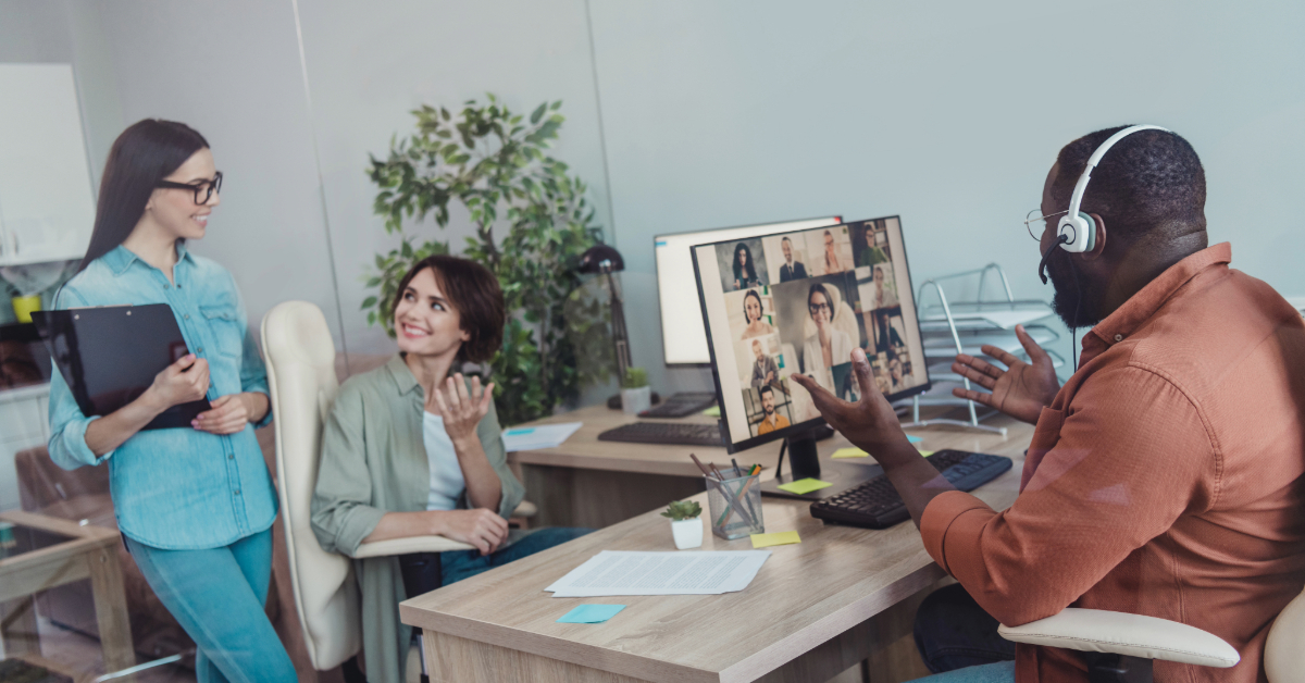 Three colleagues in an office, with one man on a video call wearing headphones, while two women stand and sit nearby, interacting and smiling as they discuss business tips.