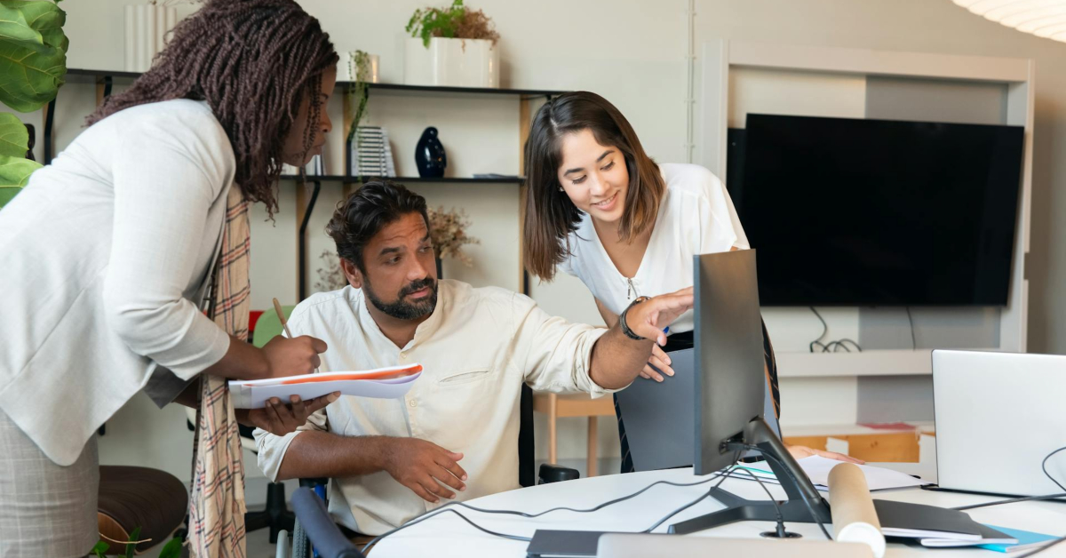 Three people collaborate at a desk, looking at a computer monitor—one gestures at the screen while another takes notes, reflecting the teamwork essential for running a successful agency in 2024.