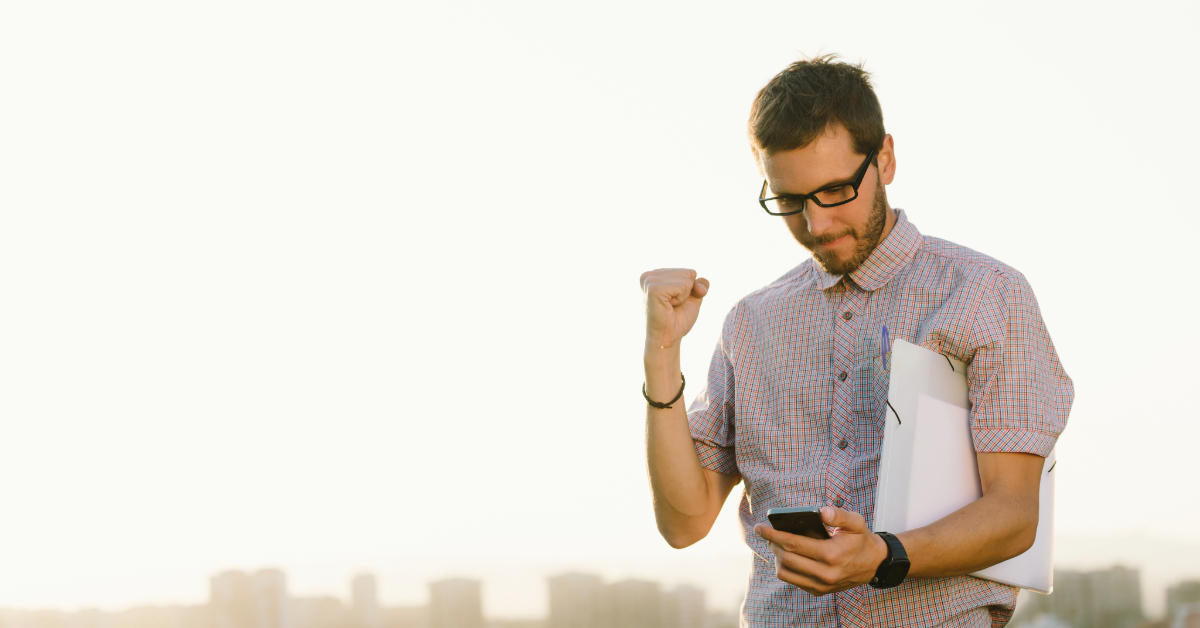 A man wearing glasses holds a laptop and looks at his phone with a fist raised, clearly pleased—perhaps celebrating entrepreneurial success—outdoors with a cityscape background.