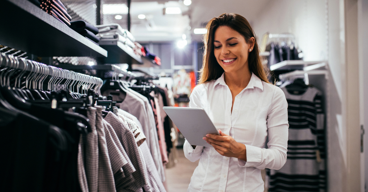 A woman in a white shirt stands in a retail store, smiling while using a tablet. Clothing racks and shelves are visible in the background, suggesting ways to increase footfall and engage more shoppers.