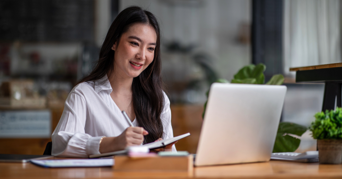 A woman sits at a desk, smiling while writing in a notebook and looking at a laptop screen in a well-lit indoor workspace, planning e-Commerce solutions.