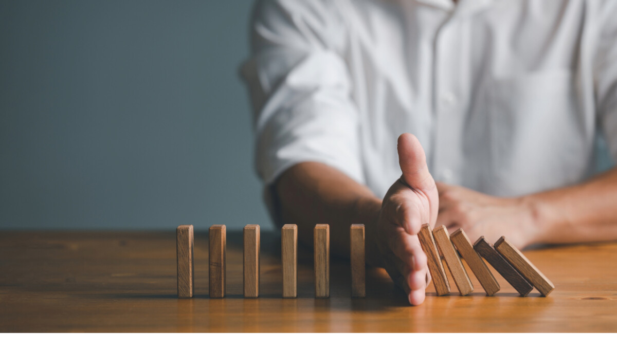 A person in a white shirt stops a row of falling wooden dominoes with their hand on a wooden table, symbolizing how political uncertainty can interrupt momentum and have serious business implications.