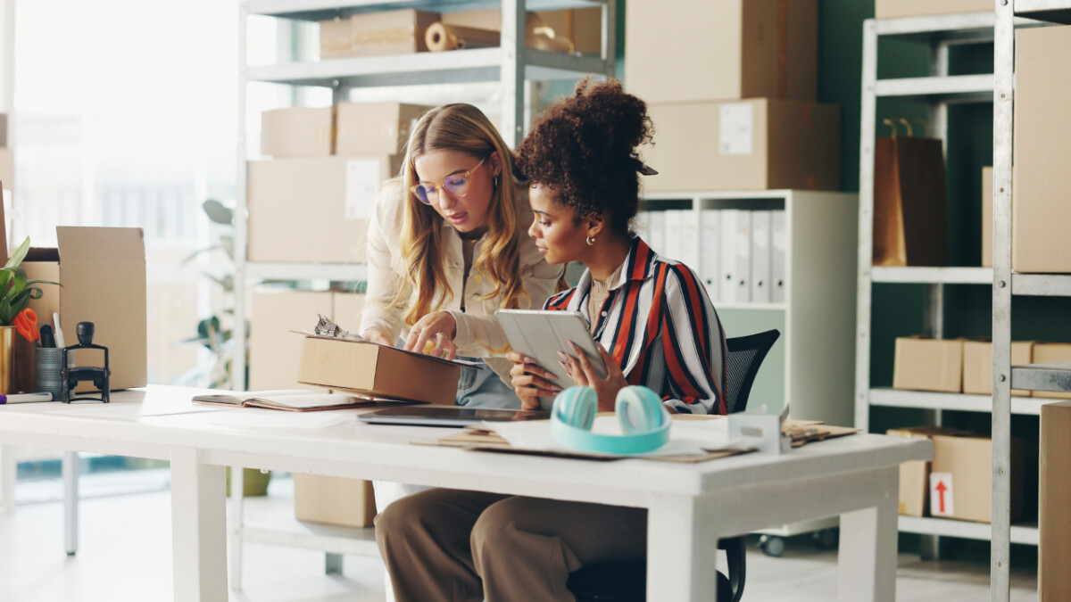 Two women sit at a desk in a workspace with shelves of boxes; one inspects items in a box while the other holds a tablet, reflecting the teamwork behind e-commerce success.