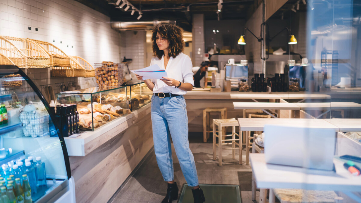 A woman stands in a modern bakery or café, holding papers and looking around, as pastries fill glass cases—perhaps reviewing franchise marketing strategies with AI-driven insights for the bustling business.