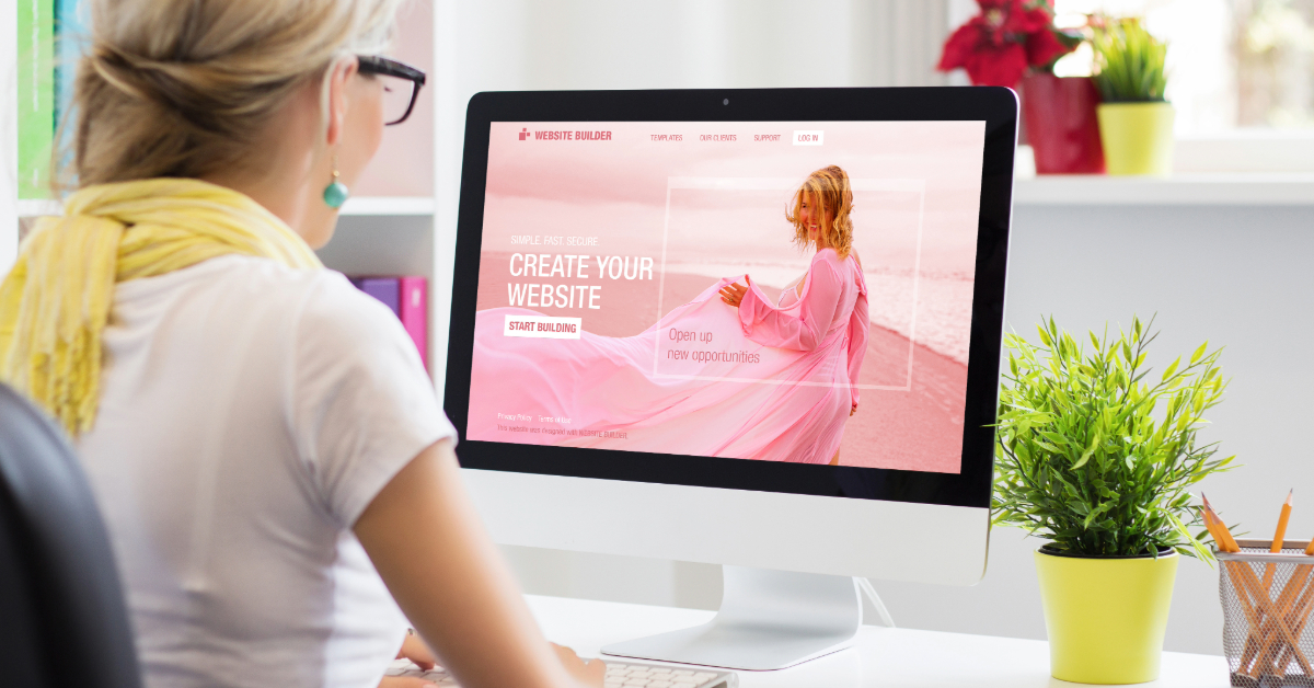 Person sitting at a desk, working on a desktop computer displaying a website builder interface with the text "Create Your Website" and featuring high-quality images of a woman in a pink dress on the screen.