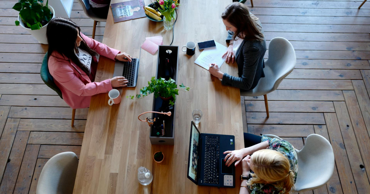 Three women are working at a wooden table with laptops, notebooks, and drinks in a modern office space, collaborating on ways to make their business stand out.