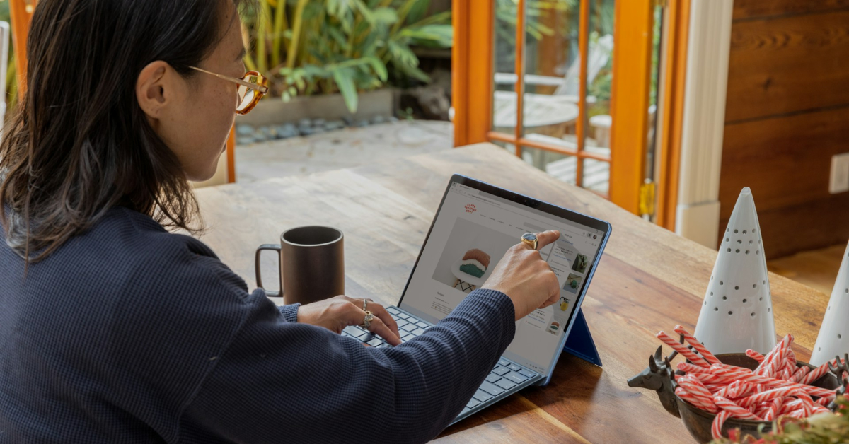 A business owner uses a stylus to interact with a touchscreen laptop at a wooden table, surrounded by a mug, holiday decorations, and plants—perhaps researching money saving tips or new techniques.