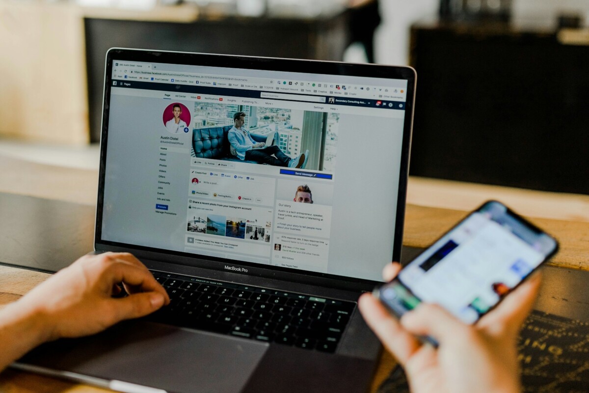 A person uses a laptop to browse a Facebook profile while holding a smartphone displaying another app—an everyday scene showcasing how small businesses can leverage Digital PR tools across multiple devices.