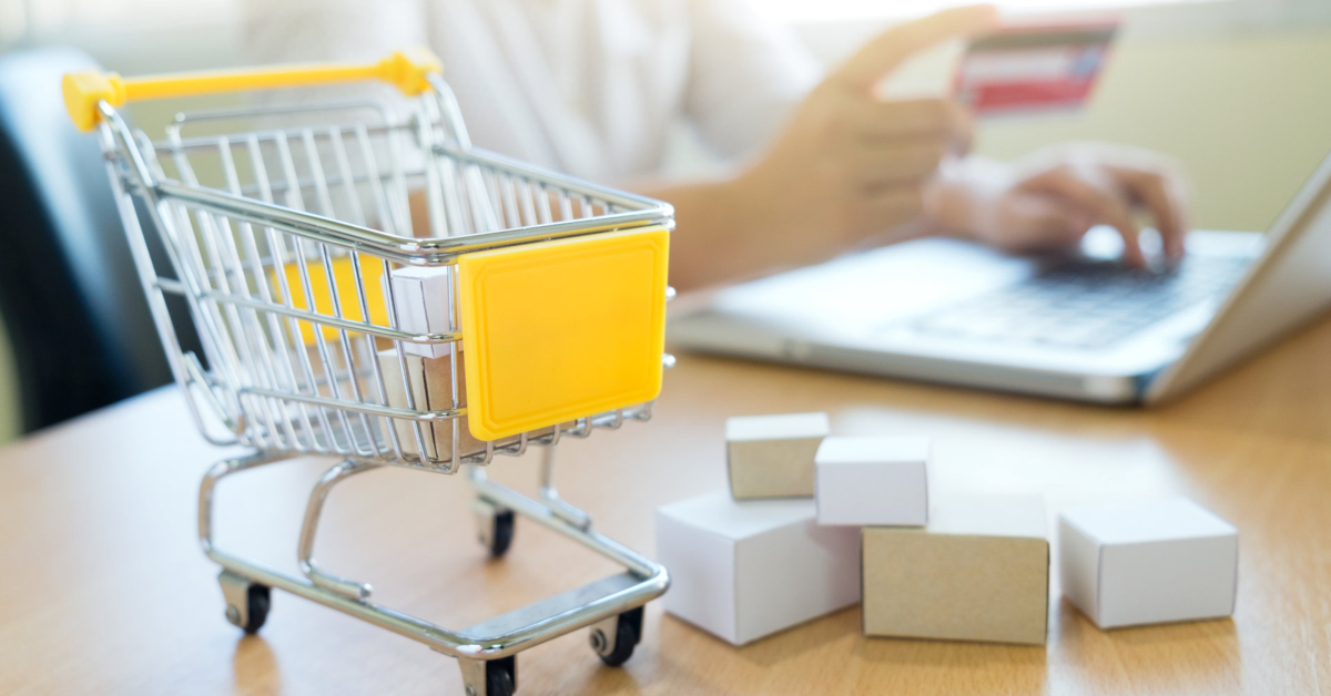 A person uses a laptop and credit card in the background, with a small shopping cart and several cardboard boxes on a table in the foreground, highlighting the impact of AI in E-Commerce on customer experience.