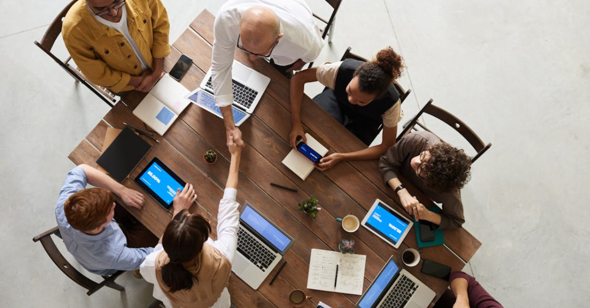 A group of people sit around a wooden table with laptops and notebooks, as two individuals shake hands across the table, discussing how to avoid common small business mistakes.