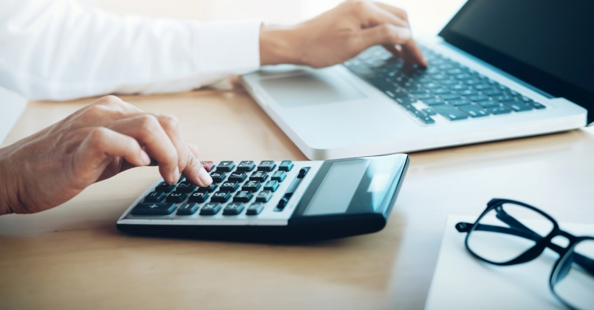 A person uses a calculator with one hand to calculate fixed asset turnover while typing on a laptop with the other; eyeglasses rest on the desk nearby.