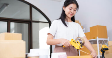 A woman seals a cardboard box with packing tape in a room filled with other boxes, reflecting the busy atmosphere of dropshipping and the importance of staying informed about industry trends.