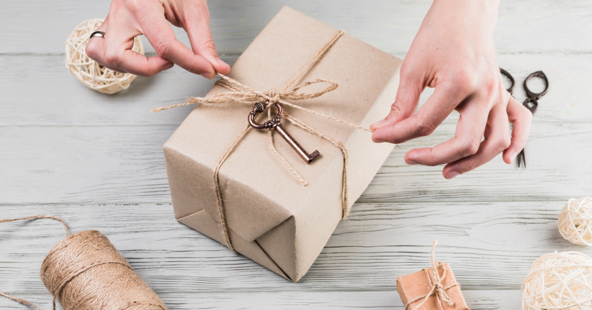 Hands tying twine around a kraft paper-wrapped gift box, decorated with a vintage key; nearby are handmade goods and wrapping materials, perfect for showcasing in online marketing shots on a rustic wooden surface.