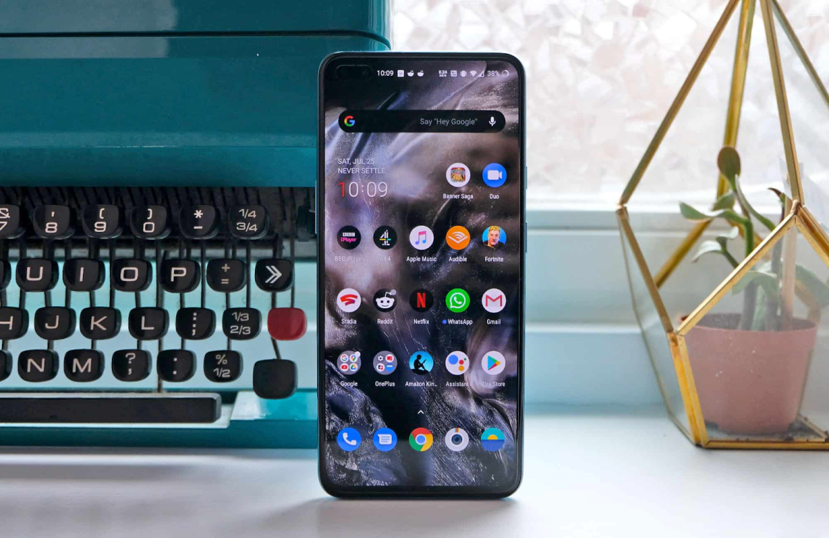 A smartphone running Android displays its home screen with various app icons, standing upright on a desk next to a vintage typewriter and a potted plant by a window—a perfect setting for iTechMoral to explore Custom ROMs.
