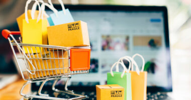 A miniature shopping cart with small boxes and bags sits in front of a laptop displaying an online shopping website, highlighting the effortless shopping experience available 24/7.