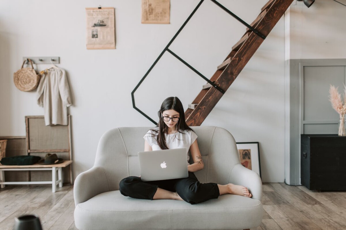 A business owner sits cross-legged on a gray sofa using a laptop in a modern living room with wooden flooring and a staircase in the background, focused on her marketing tasks.