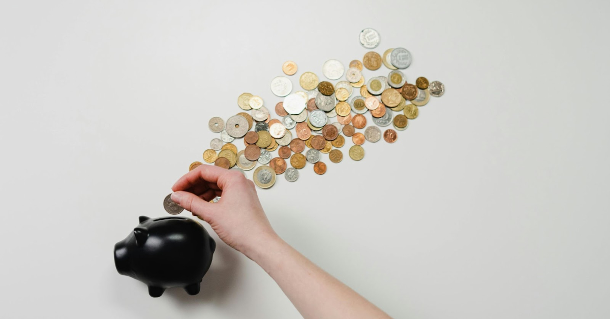 A hand places a coin into a black piggy bank, with various coins spread out on a white surface in the background—an everyday act of staying on top of finances.