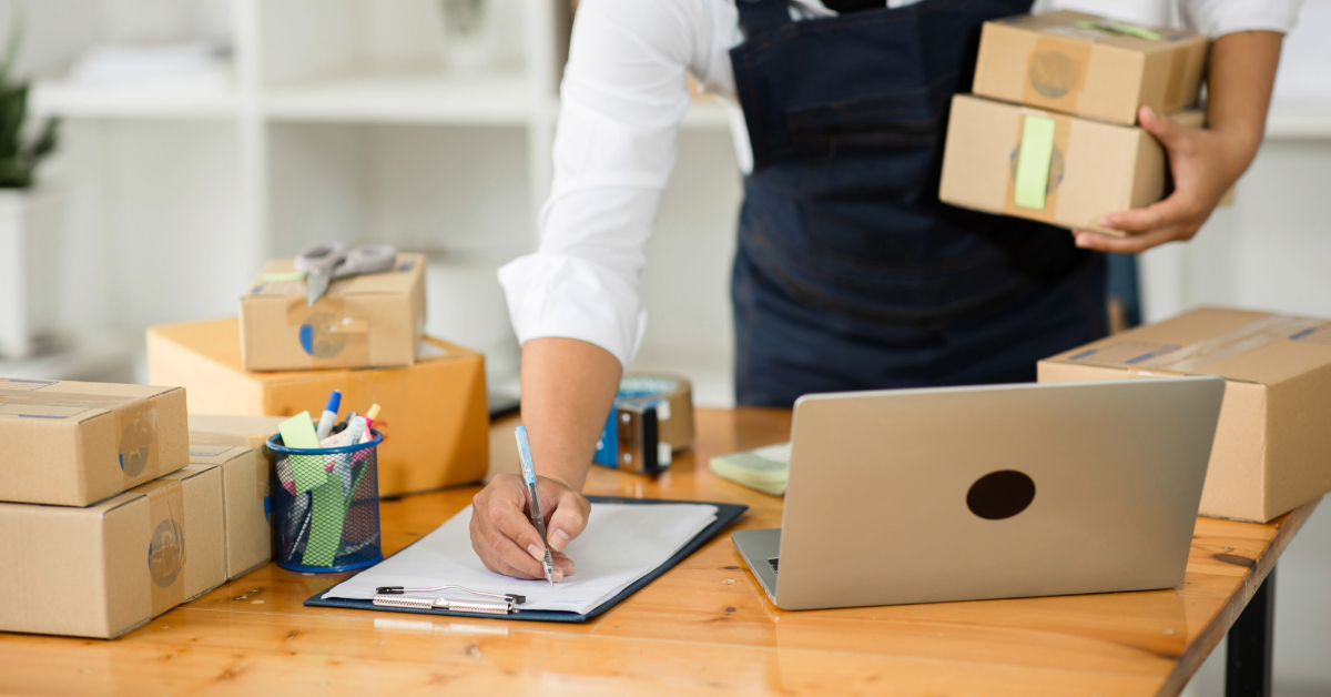 Person in an apron holding boxes and writing on a clipboard at a desk with a laptop and several packages, capturing the busy atmosphere of an ecommerce startup preparing orders for shipping.