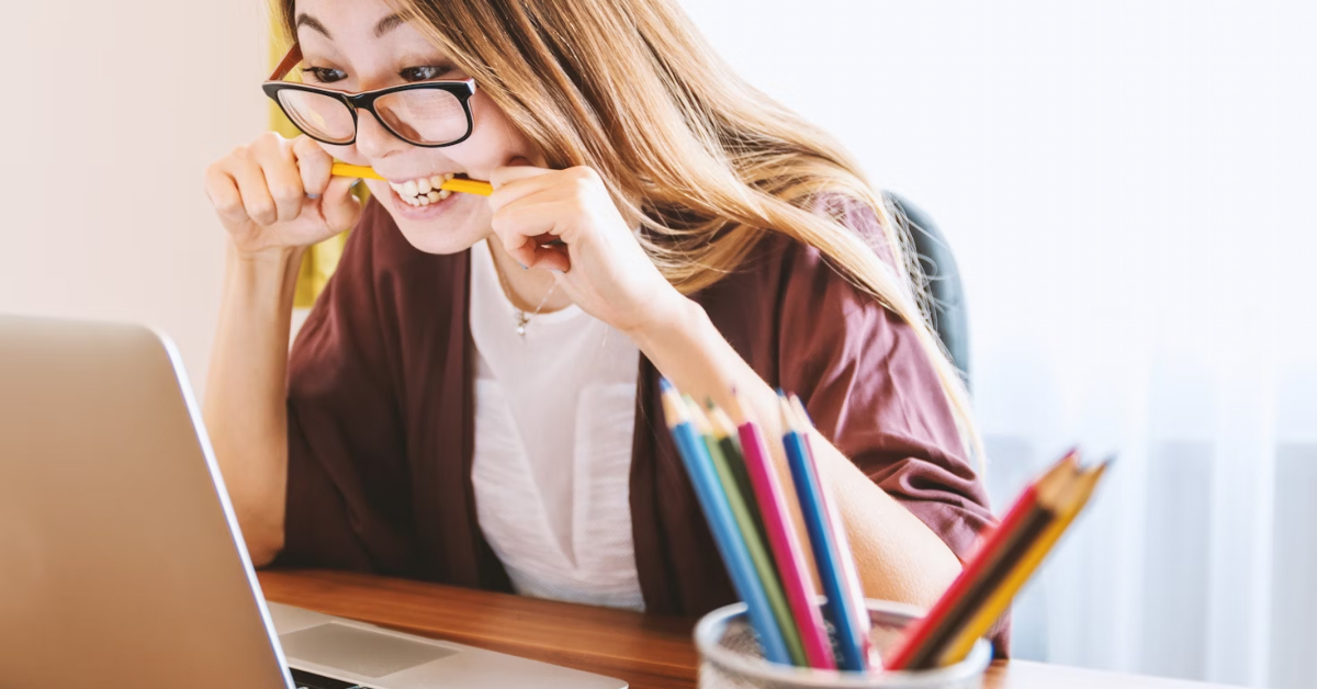 A woman sits at a desk with a laptop, biting a pencil and looking at the screen, considering strategies for improving her work environment; a cup of colored pencils sits in the foreground.