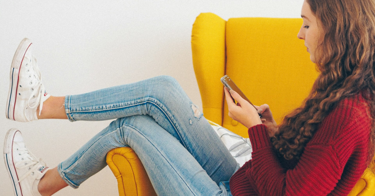 A young woman sits on a yellow chair, wearing a red sweater, jeans, and white sneakers, looking at her smartphone—possibly checking updates for her ecommerce business.