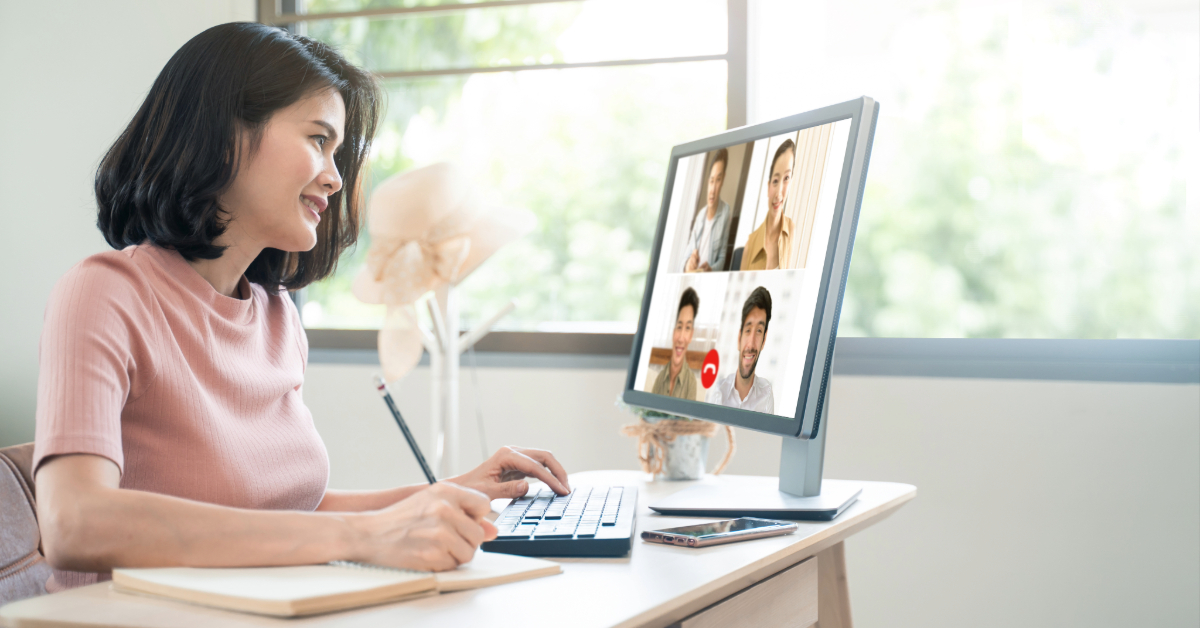 A woman sits at a desk with a notepad, participating in a video conference on her computer with four people on the screen, enhancing team collaboration through remote team engagement.