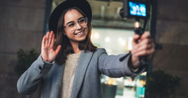 A person wearing glasses and a hat smiles and waves at a camera on a handheld tripod, appearing to record a YouTube channel video outdoors in an urban setting.