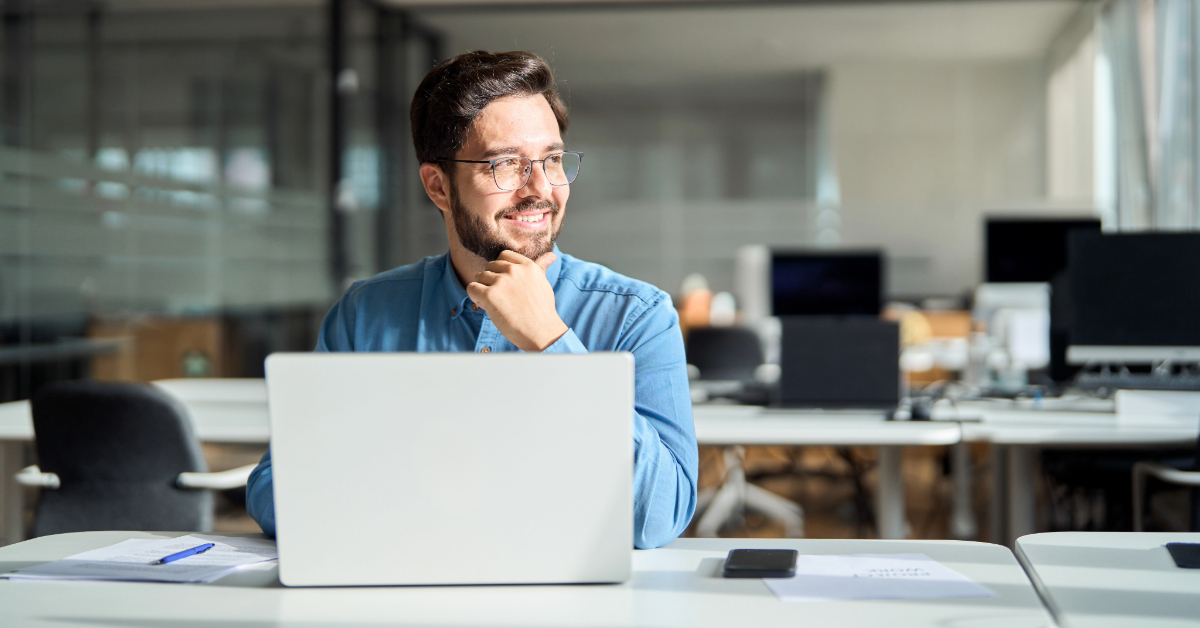 Man with beard and glasses sits at a desk in a modern office, smiling and looking to the side, with an open laptop and papers in front of him—ready to discuss business growth in 2024 using the latest startup tools.
