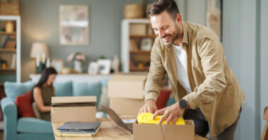 A man packing a yellow item into a cardboard box at a table, possibly preparing an eBay or Amazon order, with a woman sitting on a couch in the background.