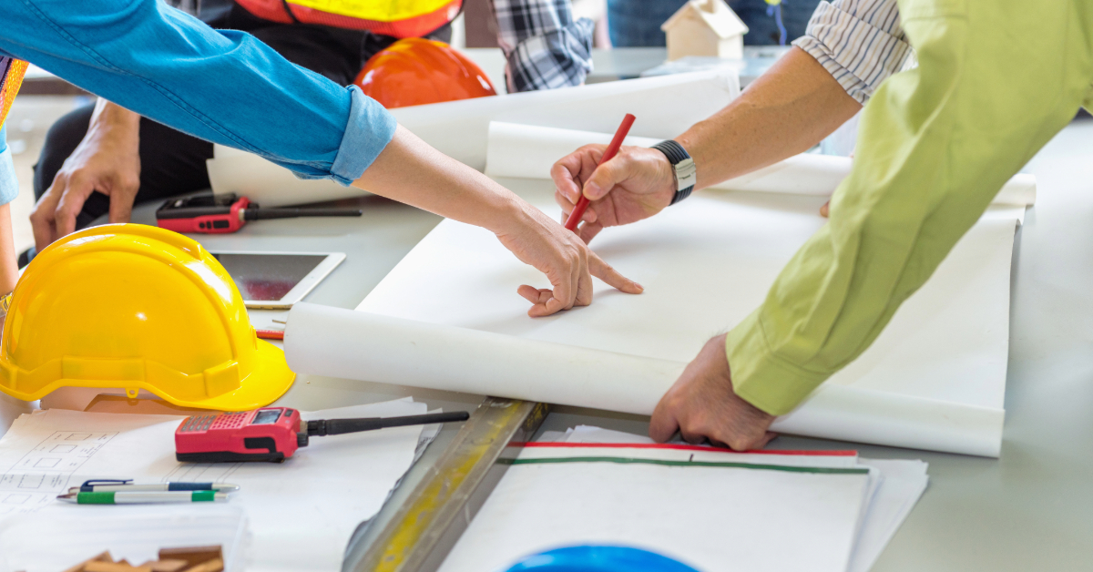Several people, wearing work shirts and safety vests, collaborate over construction blueprints on a table with hard hats and measuring tools, streamlining construction project management through organized teamwork and detailed planning.