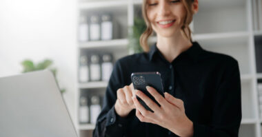 A woman in a black shirt stands by a laptop, smiling while using her smartphone—perhaps sharing her digital business card—in an office setting with shelves in the background.