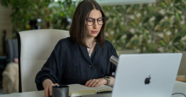A woman with glasses sits at a desk using a laptop, with a notebook, pen, and mug in front of her—possibly exploring roles in e-commerce live streaming. Leafy wallpaper and plants add charm to the background.