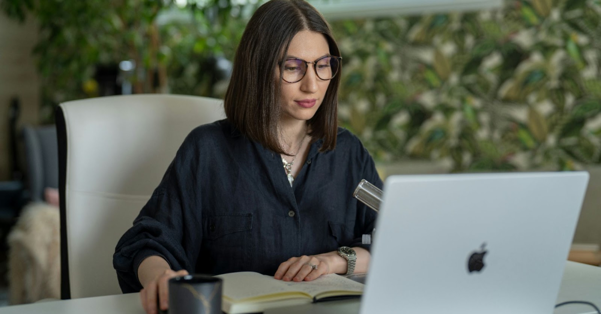 A woman with glasses sits at a desk using a laptop, with a notebook, pen, and mug in front of her—possibly exploring roles in e-commerce live streaming. Leafy wallpaper and plants add charm to the background.