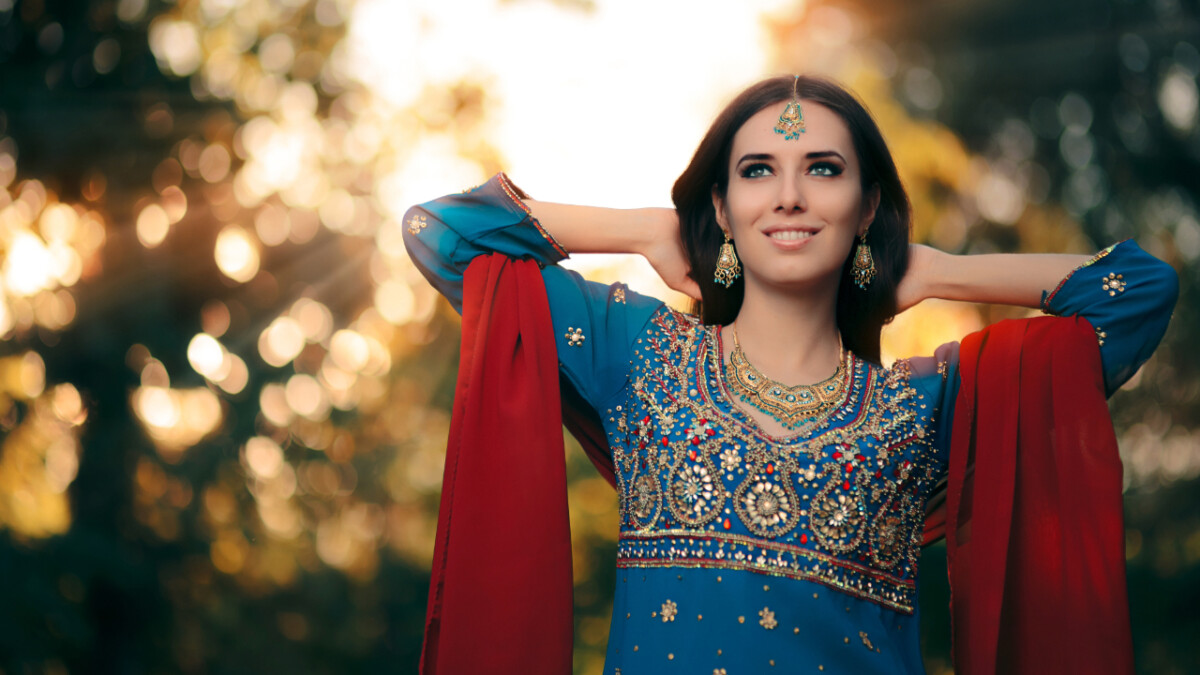 Woman in traditional blue and red embroidered attire stands outdoors with her hands behind her head, smiling brightly—just like the confidence that comes when you learn how to sell on Shopify. Sunlight and greenery blur softly in the background.