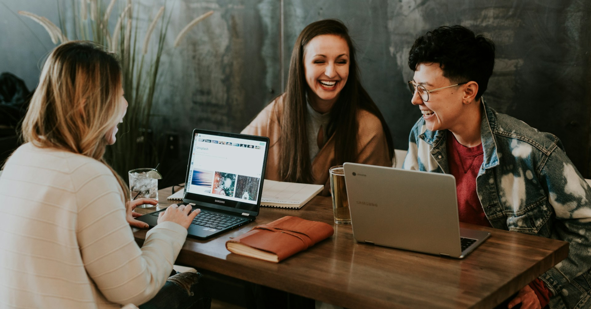 Three people sit at a wooden table using laptops and notebooks, smiling and talking together in a casual indoor setting, brainstorming sales-focused marketing strategies for e-commerce growth.