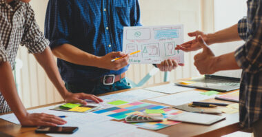 Three people review website wireframes and colorful charts on a table, with one person holding a sheet showing sketched versatile designs during a team meeting to boost customer engagement.