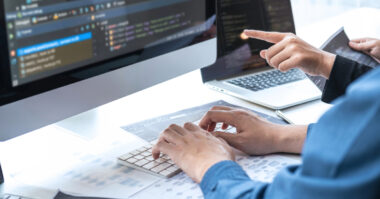 Two renewable energy software developers work at a desk with code on a computer monitor and laptop, one typing and the other pointing at the screen, discussing programming or software development.