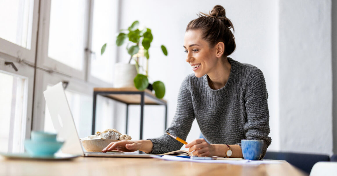 A woman sits at a desk, smiling while working on a laptop. Surrounded by notebooks, a cup, and a plant, she plans clever investments for business growth as natural light pours through large windows.