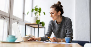A woman sits at a desk, smiling while working on a laptop. Surrounded by notebooks, a cup, and a plant, she plans clever investments for business growth as natural light pours through large windows.