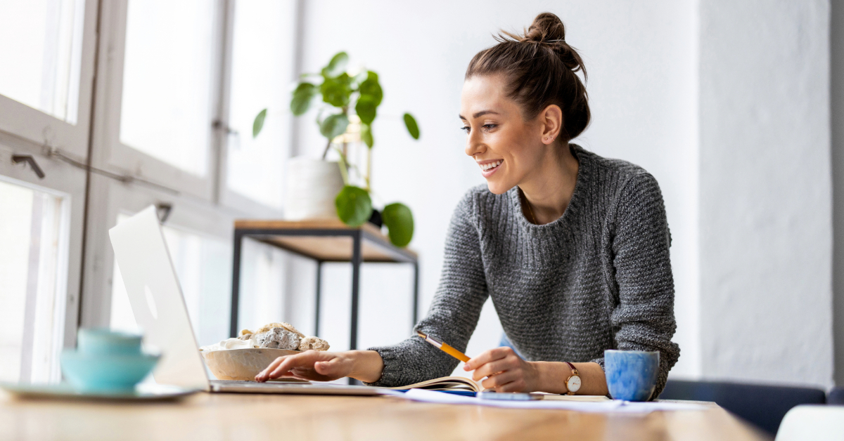 A woman sits at a desk, smiling while working on a laptop. Surrounded by notebooks, a cup, and a plant, she plans clever investments for business growth as natural light pours through large windows.